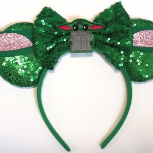 May include: Green sequined Minnie Mouse ears headband with a green bow and a character from Star Wars: The Mandalorian, Grogu, in the centre.