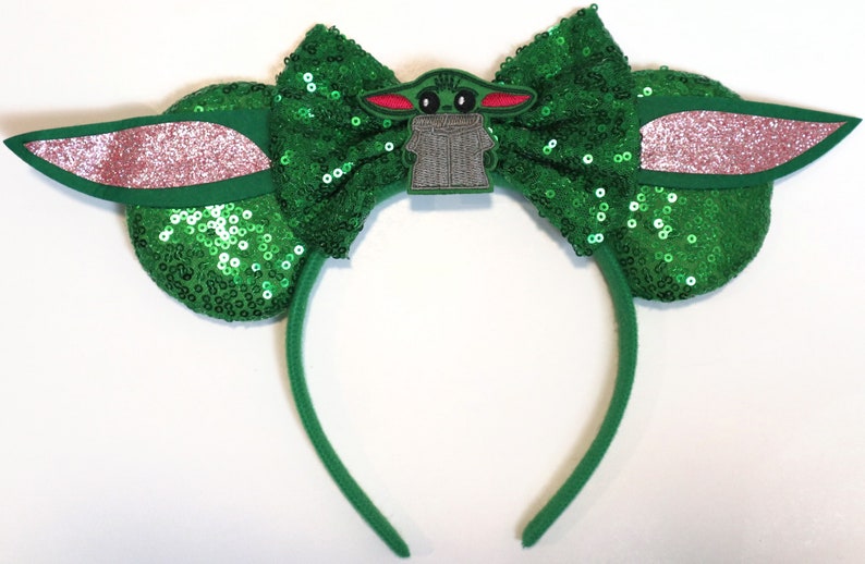 May include: Green sequined Minnie Mouse ears headband with a green bow and a character from Star Wars: The Mandalorian, Grogu, in the centre.