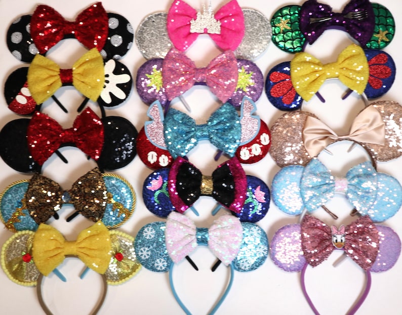 May include: A collection of 12 pairs of sparkly, colorful, sequined Minnie Mouse ears with various themed bows. The bows include a red and white polka dot bow, a yellow bow, a pink and purple bow, a blue and teal bow, a gold and brown bow, a red and blue bow, a light blue bow, a pink and white bow, a purple and pink bow, a green and purple bow, a red and yellow bow, and a gold and yellow bow.