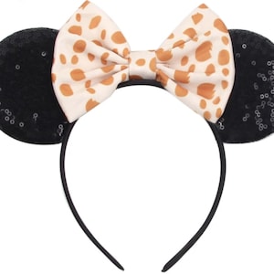 May include: A headband featuring two round black ears covered in small, shimmering sequins. Centered between the ears is a large, fabric bow in a cream colour with an irregular brown spotted pattern, resembling a giraffe or animal print design.