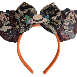 May include: A headband featuring two round ears covered in dark brown sequins. A large central bow displays a black and gold sequin pattern. A safari-themed hat sits on the bow, flanked by figures of Minnie and Mickey Mouse in safari gear.