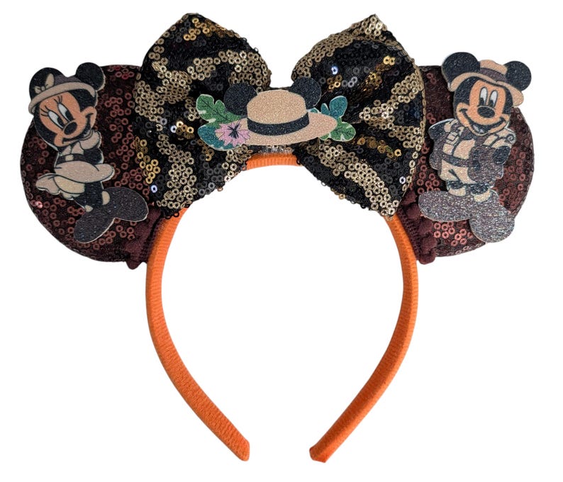 May include: A headband featuring two round ears covered in dark brown sequins. A large central bow displays a black and gold sequin pattern. A safari-themed hat sits on the bow, flanked by figures of Minnie and Mickey Mouse in safari gear.