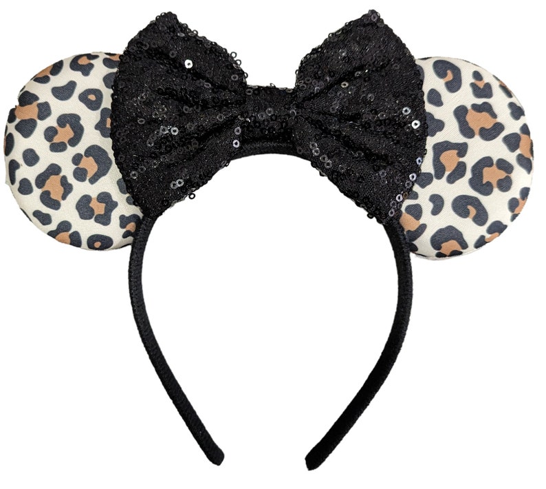 May include: A headband featuring two circular ears with a cream, tan, and black leopard print pattern. Centered between the ears is a large, upright bow covered in small, reflective black sequins. The headband frame is a solid black material.