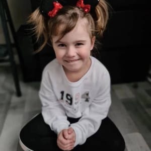 Puede incluir: Una persona joven con una camiseta blanca de manga larga con el n&uacute;mero "1994" impreso en la parte delantera. Lleva pantalones negros y una diadema negra con orejas negras y lazos rojos con lentejuelas. La persona est&aacute; sonriendo.