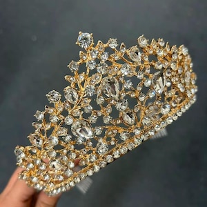 Pode incluir: Uma coroa dourada adornada com strass claros e brilhantes. A coroa apresenta um design floral com strass de v&aacute;rios tamanhos. &Eacute; segurada por uma m&atilde;o contra um fundo escuro. &Eacute; um acess&oacute;rio nupcial ou para ocasi&otilde;es especiais.