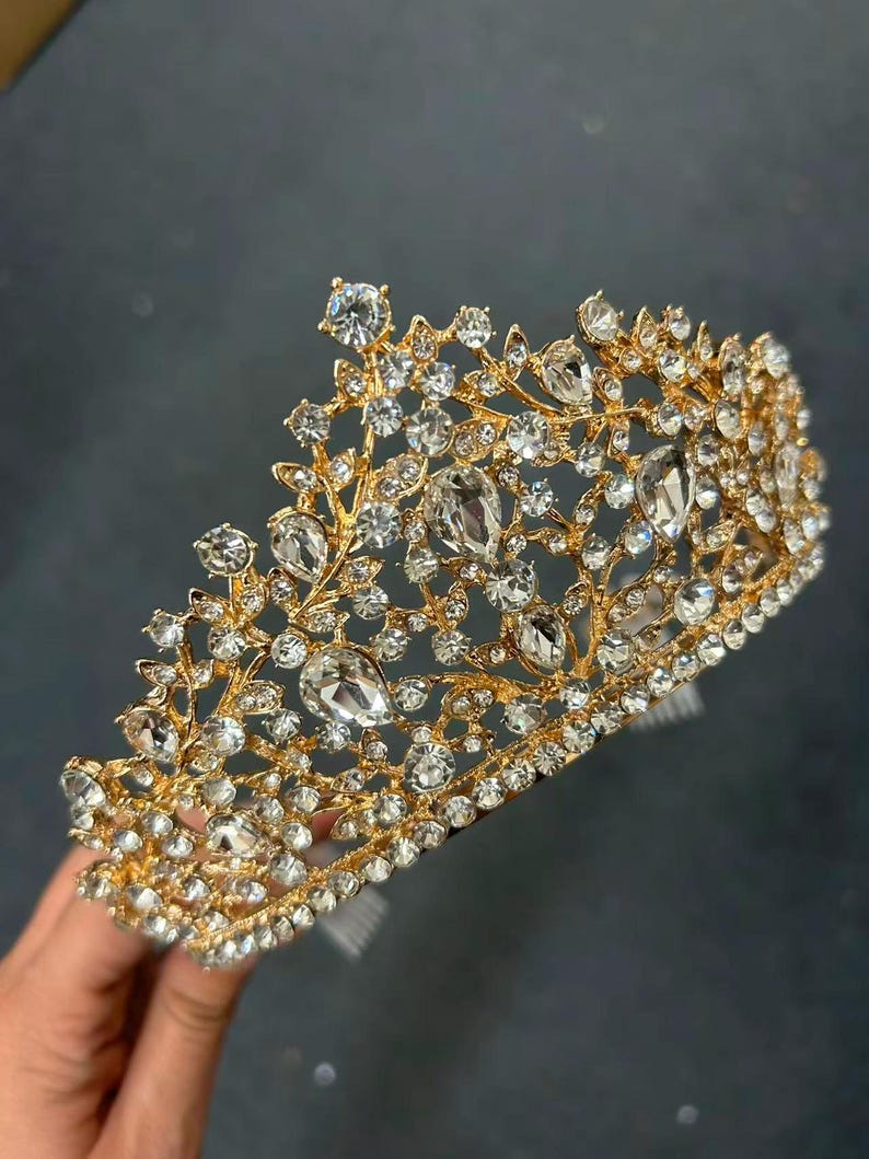 Pode incluir: Uma coroa dourada adornada com strass claros e brilhantes. A coroa apresenta um design floral com strass de v&aacute;rios tamanhos. &Eacute; segurada por uma m&atilde;o contra um fundo escuro. &Eacute; um acess&oacute;rio nupcial ou para ocasi&otilde;es especiais.