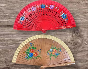 May include: Two folding hand fans are displayed. The top fan is bright red with intricate cutouts and hand-painted blue, purple, and white floral details. The bottom fan is light brown with a cream-colored top edge and painted floral clusters.