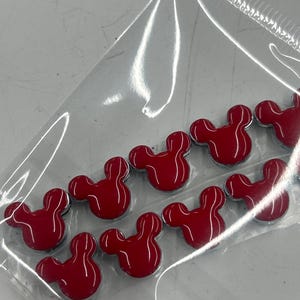 Puede incluir: Un conjunto de diez pequeños dijes de esmalte rojo brillante con forma de ratón, dispuestos en una bolsa de plástico transparente. Cada dije presenta la silueta icónica de una cabeza redonda con dos orejas y un acabado liso y reflectante.