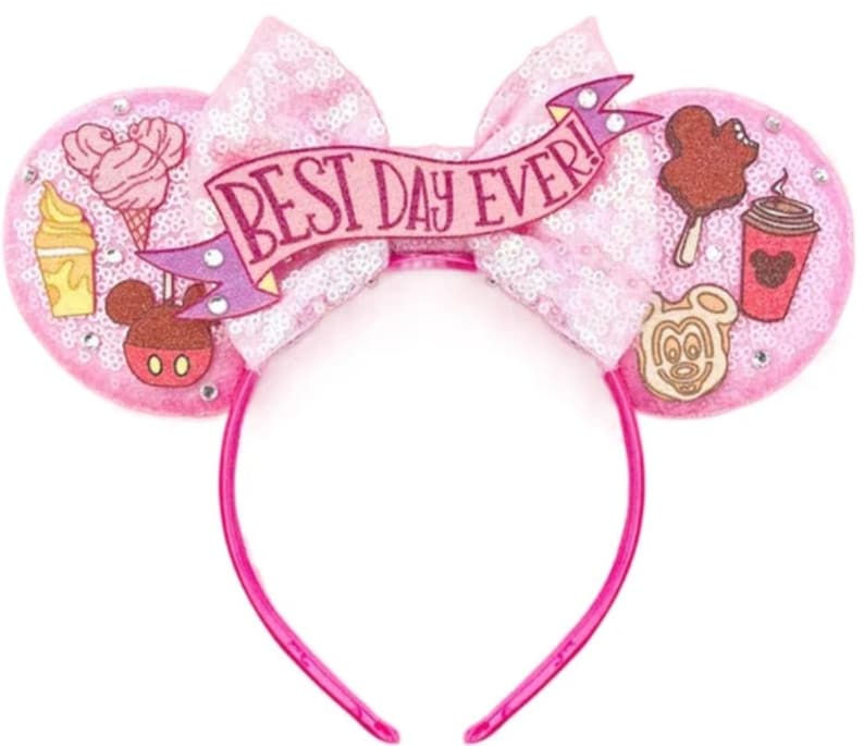 May include: A pink sequined headband featuring two circular ears and a central bow. The bow displays a banner with the text BEST DAY EVER! The ears are decorated with various food-themed icons including ice cream, coffee, and Mickey-shaped treats.