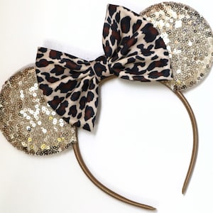 May include: A headband featuring two circular ears covered in shimmering gold sequins and a central fabric bow with a brown and black leopard print pattern. The headband frame is a solid brown colour, providing a base for the decorative elements.