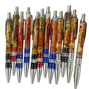 May include: A collection of ballpoint pens featuring religious imagery. The pens have silver tips and tops, with decorative bands in red, blue, and black. The pen barrels display colorful illustrations of religious figures.