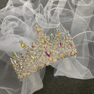 Pode incluir: Uma coroa prateada com pedras preciosas iridescentes, repousando sobre um v&eacute;u de tule branco. A coroa apresenta um design de filigrana detalhado e uma variedade de pedras coloridas e brilhantes. O v&eacute;u &eacute; transparente e delicado.