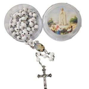 May include: A white rosary with a silver crucifix and connecting chain. The rosary beads are in a clear plastic container, with an image of a religious figure on the lid. The rosary is displayed on a white background.