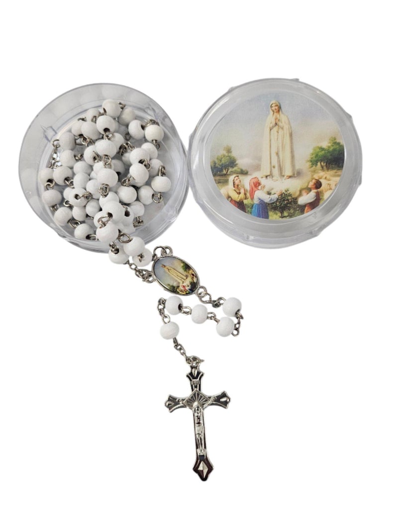 May include: A white rosary with a silver crucifix and connecting chain. The rosary beads are in a clear plastic container, with an image of a religious figure on the lid. The rosary is displayed on a white background.
