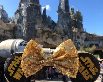 Puede incluir: Orejas de ratón negras con un lazo de lentejuelas doradas y el texto "Star Wars" en cada oreja. Las orejas se sostienen frente a un paisaje rocoso.