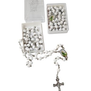 May include: White beaded rosary with a silver-tone crucifix and a small oval image. The rosary is displayed with two open white boxes filled with beads and a clear plastic box with a saint illustration.