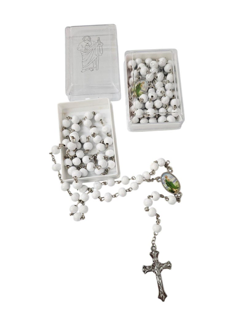 May include: White beaded rosary with a silver-tone crucifix and a small oval image. The rosary is displayed with two open white boxes filled with beads and a clear plastic box with a saint illustration.