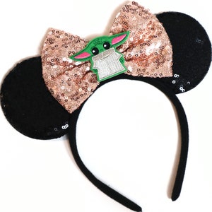 May include: Black mouse ear headband with a rose gold sequin bow and a green character patch. The character is wearing a brown robe and has large eyes.