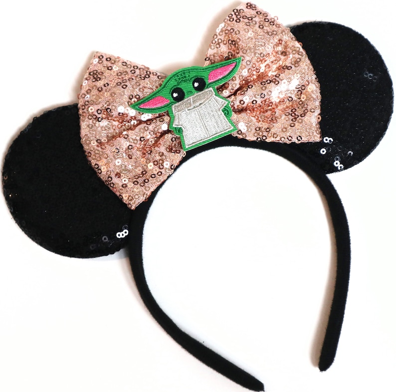May include: Black mouse ear headband with a rose gold sequin bow and a green character patch. The character is wearing a brown robe and has large eyes.