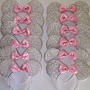 May include: Several silver glitter mouse ear headbands with pink sequin bows. The ears are round and sparkly, and the bows are a bright pink color. The headbands are white and appear to be made of metal.