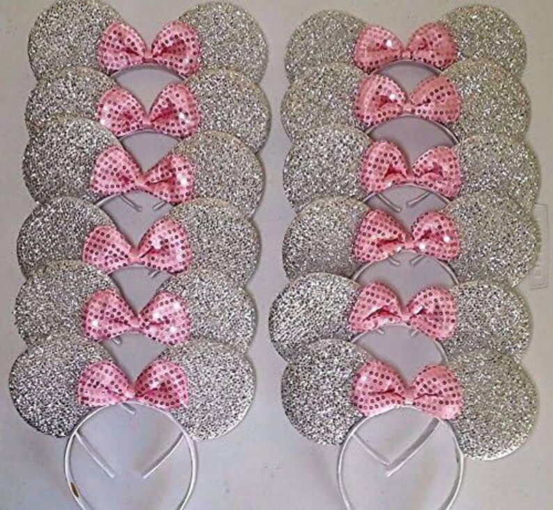 May include: Several silver glitter mouse ear headbands with pink sequin bows. The ears are round and sparkly, and the bows are a bright pink color. The headbands are white and appear to be made of metal.