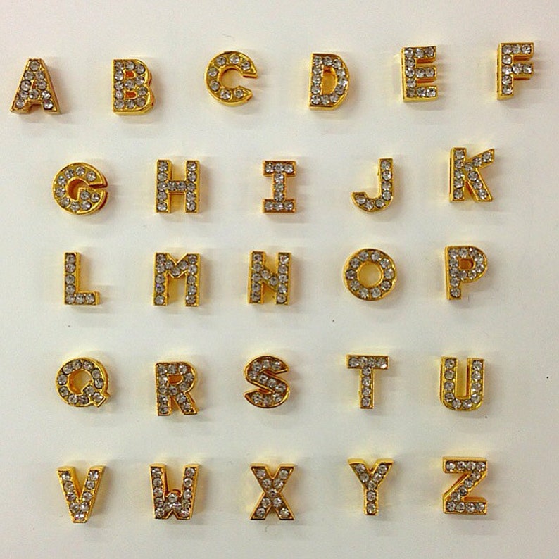 May include: Gold-toned alphabet letter charms with clear rhinestone embellishments. Each letter is individually displayed on a white background.