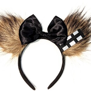 May include: Black headband with a large black satin bow and two brown furry ears. One ear has a black and white chequered ribbon.