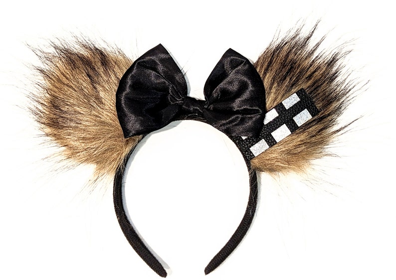 May include: Black headband with a large black satin bow and two brown furry ears. One ear has a black and white chequered ribbon.