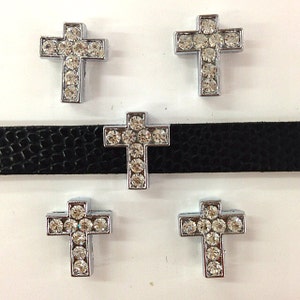 May include: Four silver cross charms with clear rhinestones. The charms are on a black leather background.