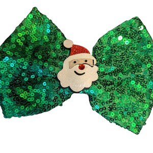 Può includere: Fiocco di paillettes verde con un viso di Babbo Natale bianco al centro. Il viso di Babbo Natale ha un cappello rosso e una barba bianca.