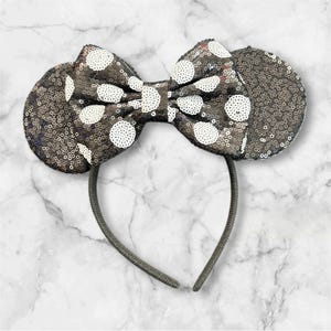 May include: A headband featuring two circular ears and a central bow, all covered in dark grey sequins. The bow is accented with white sequined polka dots. The headband frame is a thin, dark grey band that connects the ears and the bow together.
