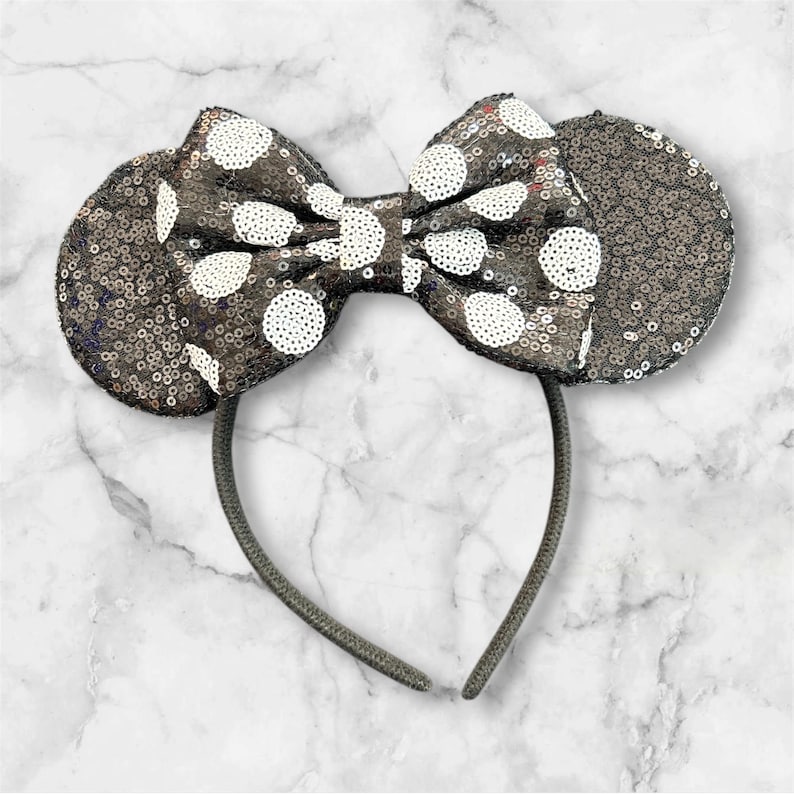 May include: A headband featuring two circular ears and a central bow, all covered in dark grey sequins. The bow is accented with white sequined polka dots. The headband frame is a thin, dark grey band that connects the ears and the bow together.