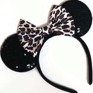May include: A headband featuring two round black ears covered in small, reflective sequins. Centered between the ears is a large, fabric bow with a beige, brown, and black leopard print pattern. The headband frame is wrapped in a soft black fabric.