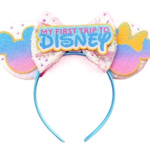 May include: A headband featuring two circular ears with a blue, pink, and purple gradient finish. A large, light pink sequined bow sits in the centre, topped with a patch that reads MY FIRST TRIP TO DISNEY in blue and pink lettering.