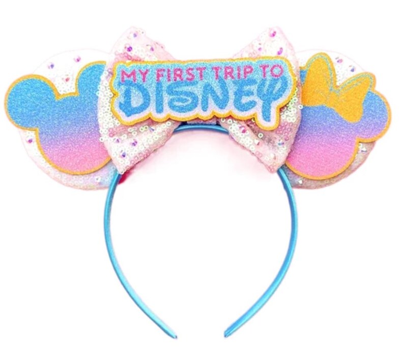 May include: A headband featuring two circular ears with a blue, pink, and purple gradient finish. A large, light pink sequined bow sits in the centre, topped with a patch that reads MY FIRST TRIP TO DISNEY in blue and pink lettering.