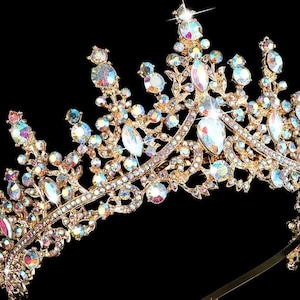 Pode incluir: Uma tiara dourada com strass iridescentes e cristais transparentes em forma de gota. A coroa ornamentada apresenta um design de filigrana detalhado sobre um fundo preto. Um acess&oacute;rio glamouroso para ocasi&otilde;es especiais.