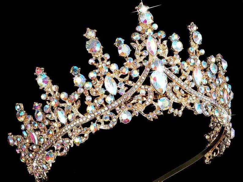 Pode incluir: Uma tiara dourada com strass iridescentes e cristais transparentes em forma de gota. A coroa ornamentada apresenta um design de filigrana detalhado sobre um fundo preto. Um acess&oacute;rio glamouroso para ocasi&otilde;es especiais.