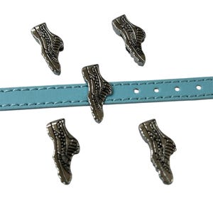 May include: Silver-colored sneaker charms are arranged around a light blue leather strap. The charms are detailed with laces and soles. The strap has evenly spaced holes. The background is white.