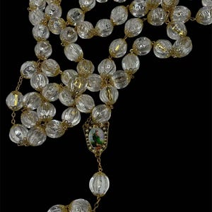 May include: A rosary featuring clear, ribbed, spherical beads separated by gold-toned metal filigree caps. The chain connects to a central shield-shaped medal depicting a figure in green robes against a landscape, finished with a gold-toned metal chain.