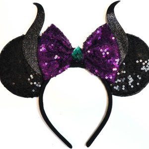 May include: Black and purple sequined headband with a large bow and a green gem in the centre. The headband has two black ears with horns.