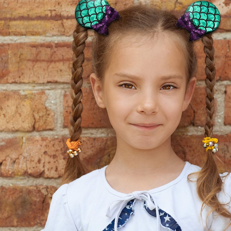 Puede incluir: Una ni&ntilde;a con cabello casta&ntilde;o largo y trenzado, adornado con pinzas para el cabello con tem&aacute;tica de sirena en verde azulado y morado. Lleva una blusa blanca con un lazo azul y blanco con estampado de estrellas. El fondo es una pared de ladrillos.