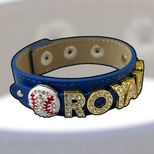 May include: Blue leather bracelet with the word "ROYALS" spelled out in gold letters with rhinestones. A baseball with red stitching and rhinestones is on the left side. The bracelet has a beige interior and silver snaps.