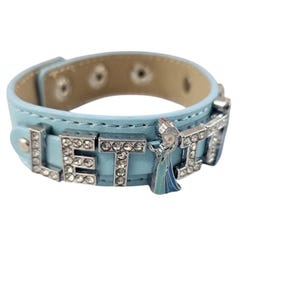 Pode incluir: Uma pulseira de couro azul claro com as palavras "LET IT" escritas em letras prateadas com strass. Um pequeno charme prateado de um personagem de desenho animado está preso à pulseira.