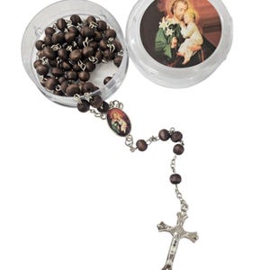 May include: A rosary with dark brown wooden beads and a silver-colored crucifix. The rosary is partially displayed inside a clear, round container. The container also features an image of a religious figure. The rosary is a religious item.