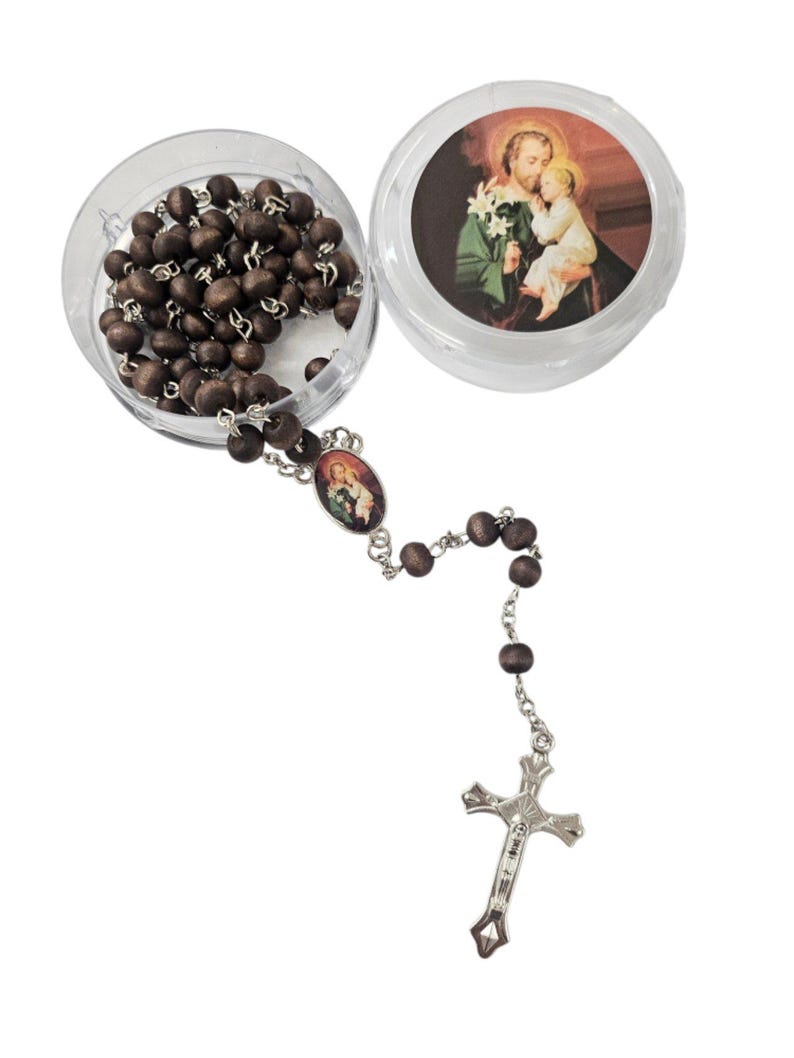 May include: A rosary with dark brown wooden beads and a silver-colored crucifix. The rosary is partially displayed inside a clear, round container. The container also features an image of a religious figure. The rosary is a religious item.