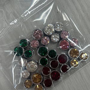 Puede incluir: Una colección de pequeños diamantes de imitación redondos y facetados en varios colores, incluyendo transparente, rosa, verde, rojo y dorado, cada uno engastado en una base de metal plateado. Estos adornos decorativos están contenidos en una bolsa de plástico transparente.