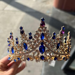 Pode incluir: Uma coroa dourada com detalhes ornamentados, com grandes pedras preciosas azuis escuras e pedras menores roxas. A coroa &eacute; adornada com cristais claros e brilhantes, criando uma apar&ecirc;ncia r&eacute;gia e elegante. O design &eacute; sim&eacute;trico.