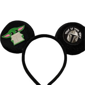 May include: Black headband with two plush ears. One ear features a green and pink embroidered character, the other ear features a black and white embroidered Mandalorian helmet with the text "This is the way".