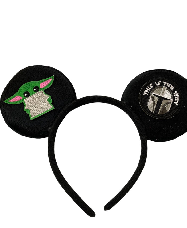 May include: Black headband with two plush ears. One ear features a green and pink embroidered character, the other ear features a black and white embroidered Mandalorian helmet with the text "This is the way".