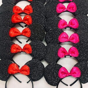 May include: Black sequined mouse ear headbands with red sequin bows and pink sequin bows. The ears are round and attached to a black headband. The bows are centered on the ears.